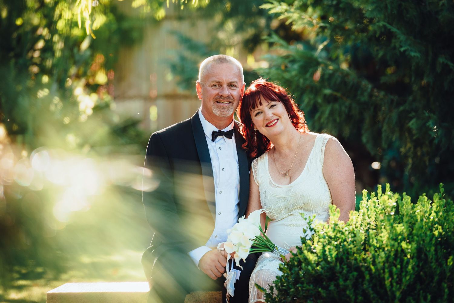 Elegant couple in formal attire smiling together outdoors in a garden setting with beautiful golden sunlight.