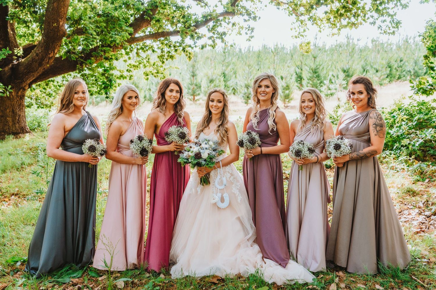 Wedding party in mismatched floor-length dresses in muted tones stand together outdoors under green trees.