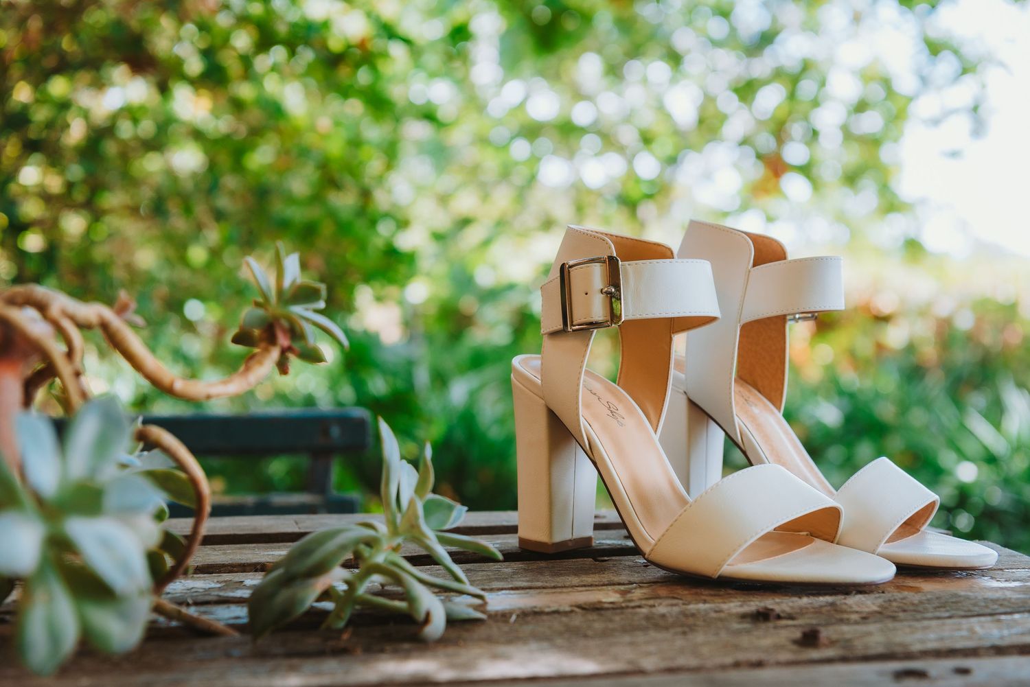 A pair of beige strappy sandals with block heels sits on a wooden surface with blurred greenery in the background.