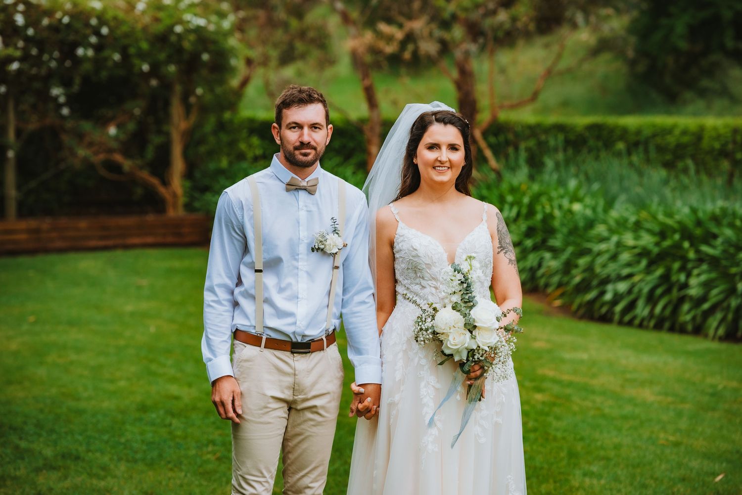 A bride and groom stand together on a lush green lawn, with the bride holding a white floral bouquet.