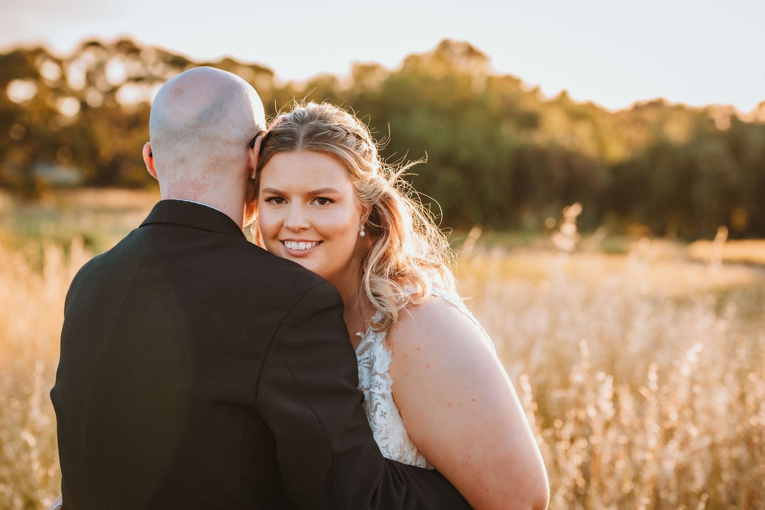 A couple embraces in a golden field at sunset as sunlight illuminates tall grass and creates a warm romantic glow.
