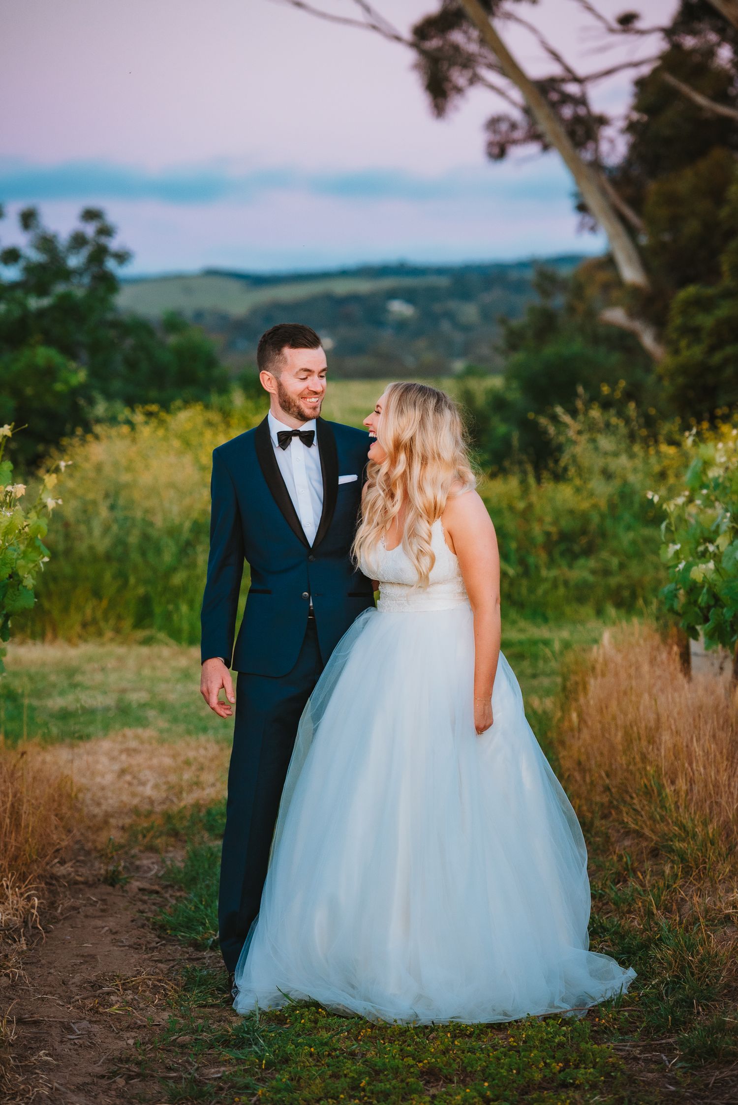 A bride in a flowy white gown and groom in navy tuxedo pose together on a hilltop during golden hour.