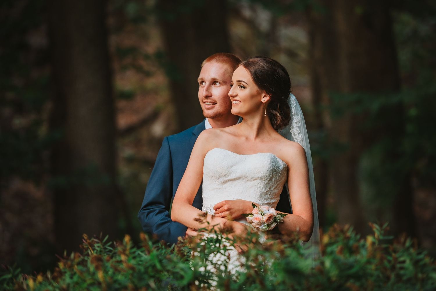 A couple in formal wedding attire embrace in a romantic forest setting with soft glowing light.