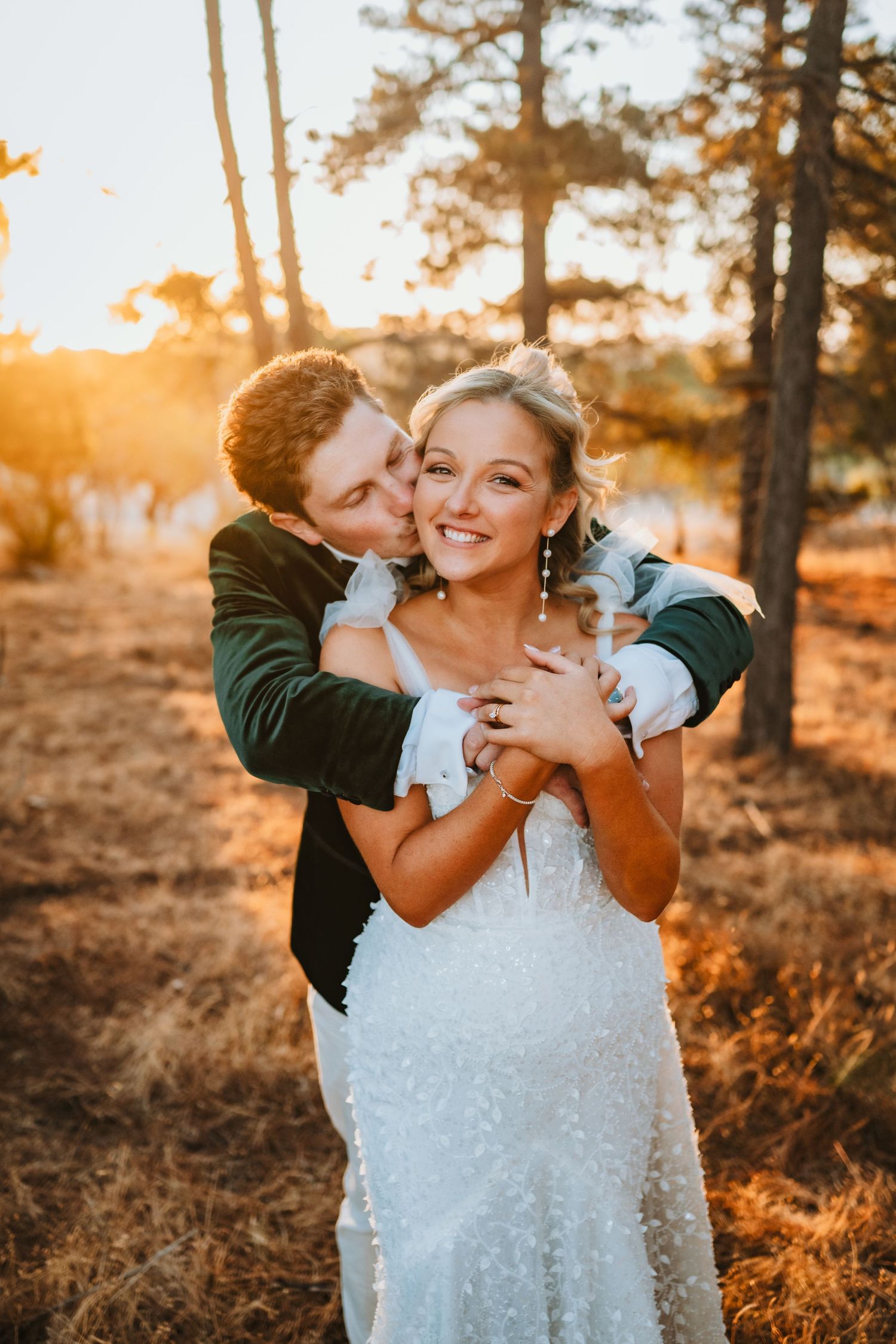 A romantic wedding photo of a couple embracing in a forest at golden sunset hour, with warm glowing backlighting.
