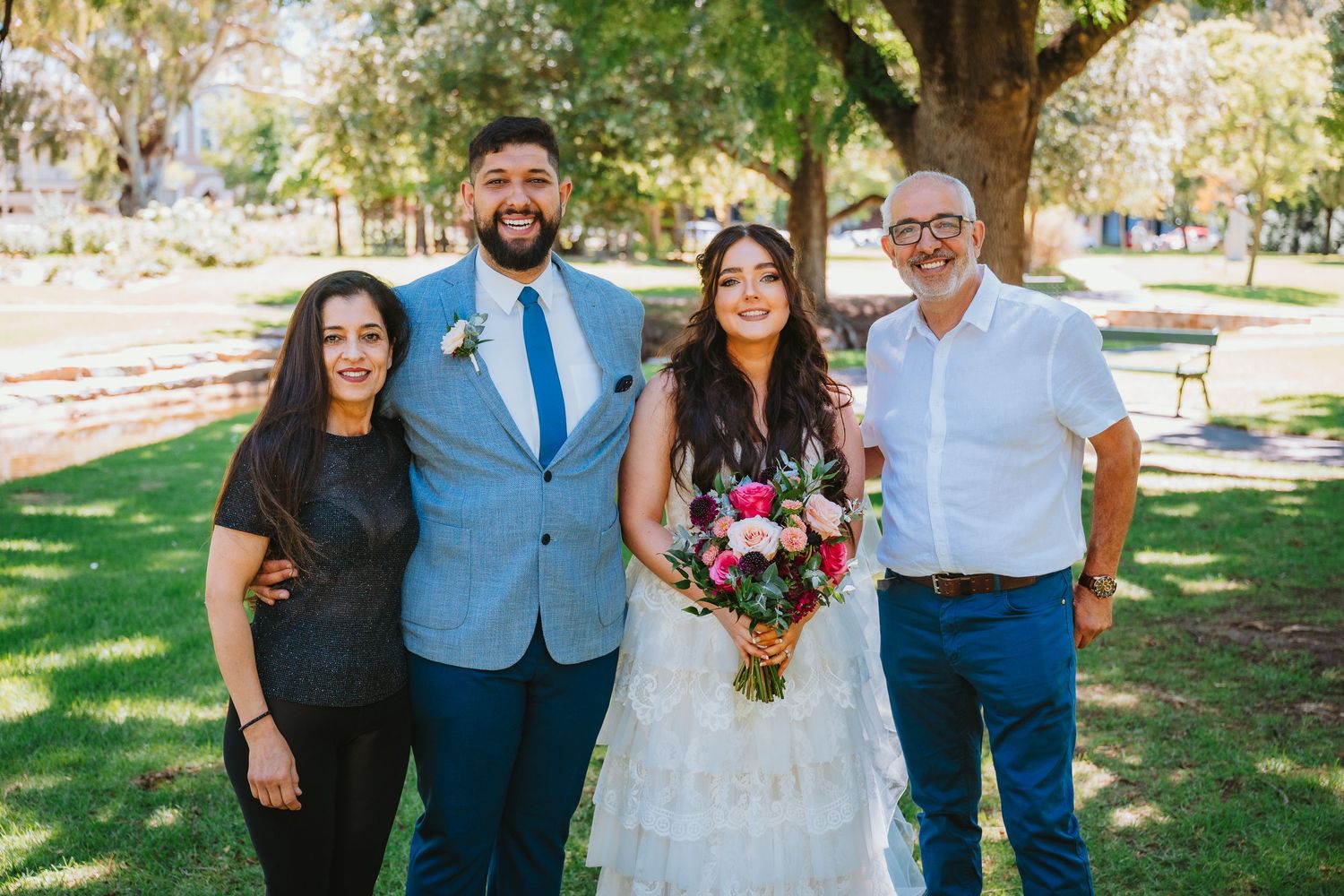 A small wedding party poses together outdoors on a sunny day in a grassy park setting with trees in the background.