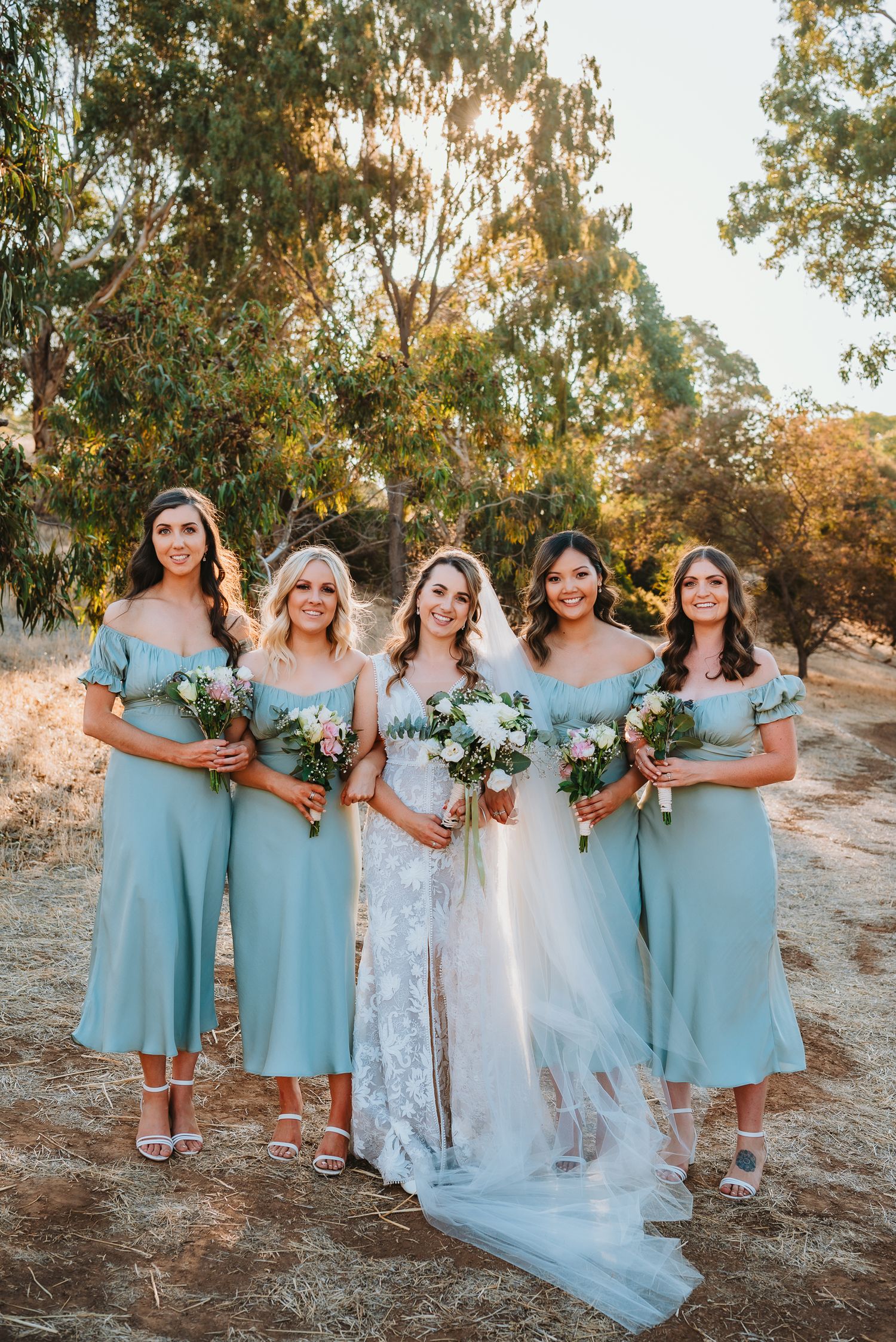 A bridal party stands together outdoors in matching powder blue dresses holding white floral bouquets at sunset.