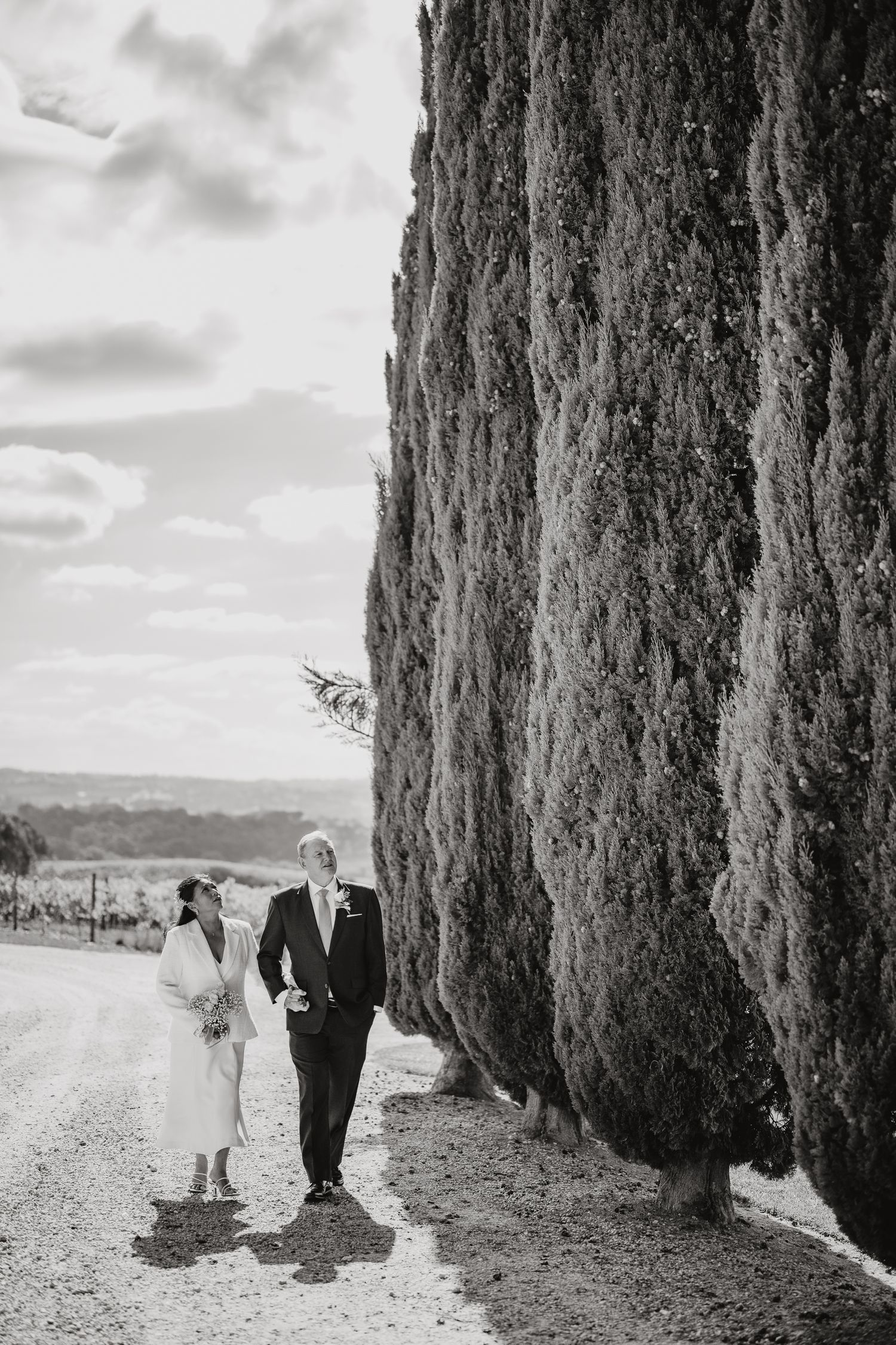 Elegant black and white photograph of a couple walking along a path beside tall cypress trees in Tuscany.