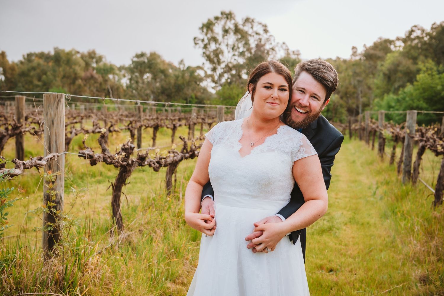 A couple embraces in a scenic vineyard with rows of grapevines stretching into the distance on a sunny day.