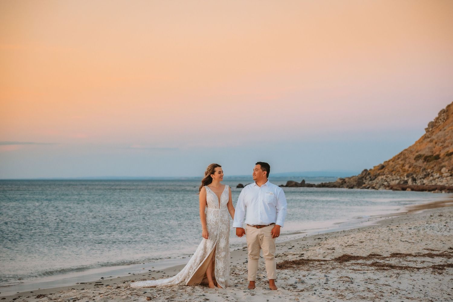 A couple in white attire walks along a beach at sunset with cliffs and ocean in the background.