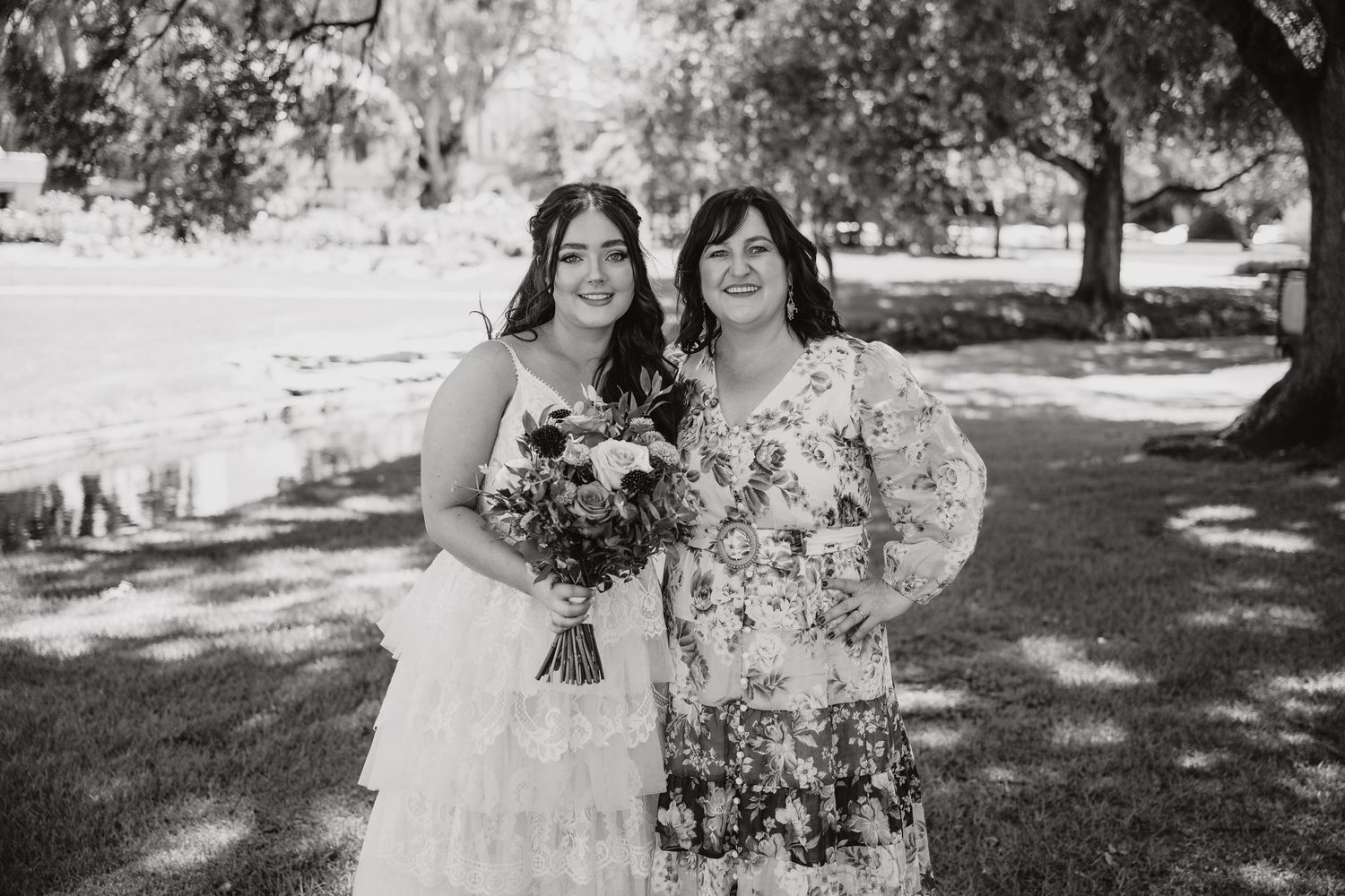 Black and white photo of two people holding floral bouquets outdoors at a sunny wedding celebration.
