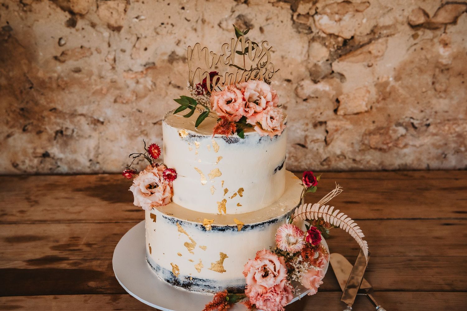 A rustic two-tier white wedding cake decorated with pink flowers and gold accents sits on a wooden table.