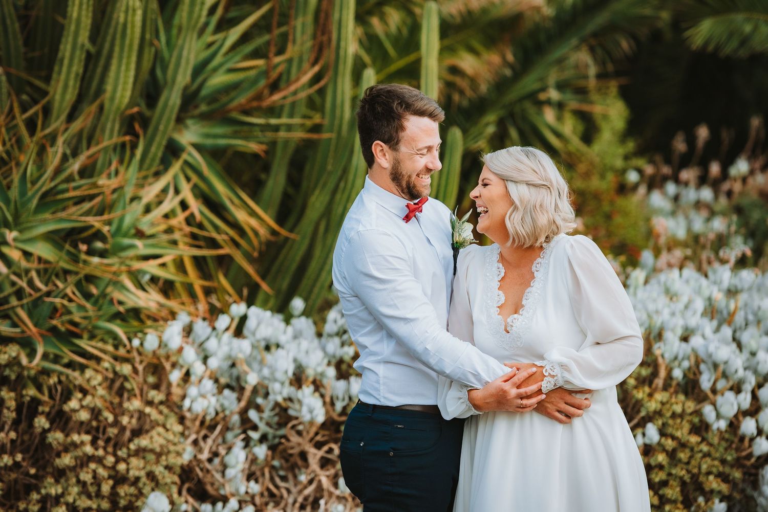 A couple shares a romantic moment in a garden surrounded by white flowers and tropical plants on their wedding day.