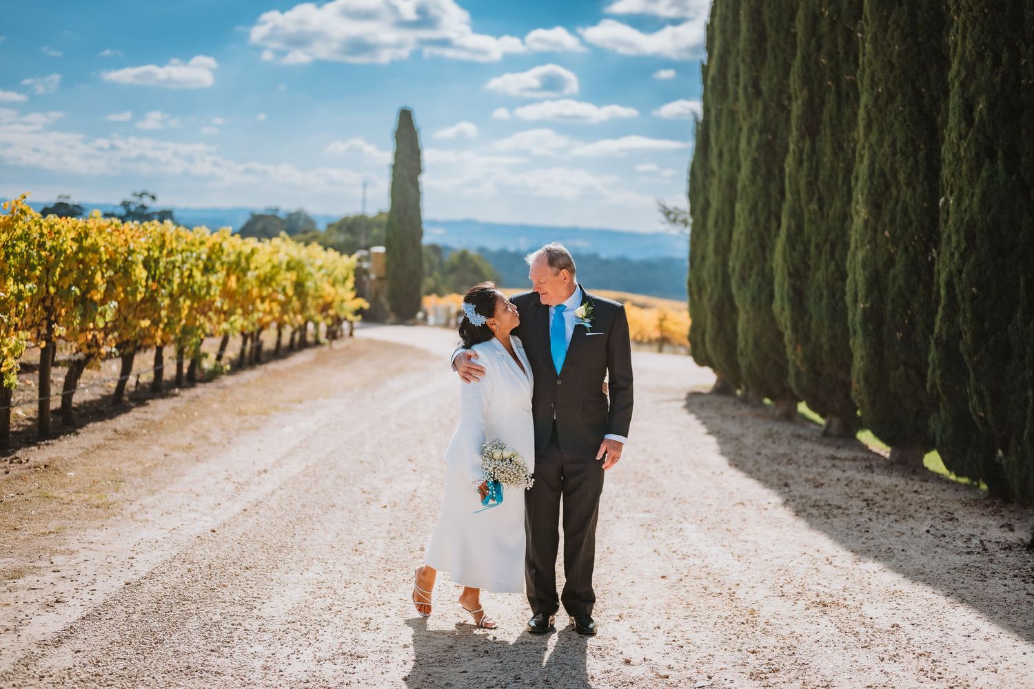 A couple walks along a scenic vineyard path lined with tall cypress trees and yellow grapevines on a sunny day.