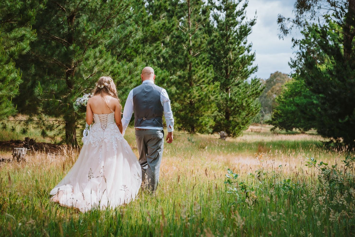 Two people walking through a grassy meadow on their wedding day with evergreen trees in the background.