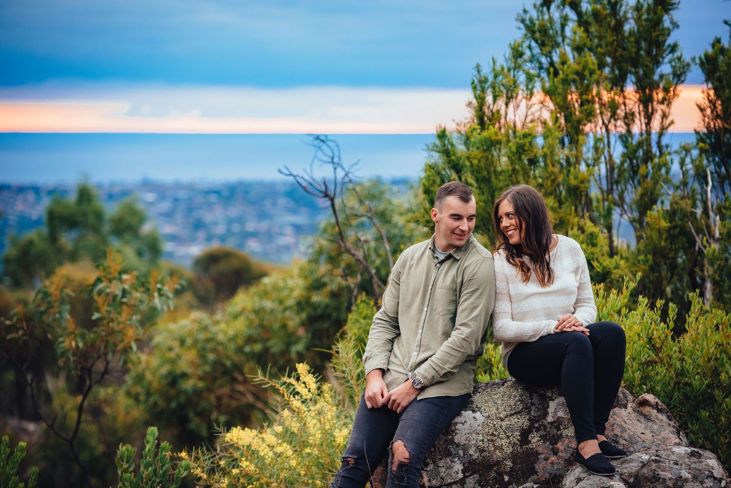 Two people sitting together on a rocky outcrop overlooking a scenic mountain landscape on a sunny day.