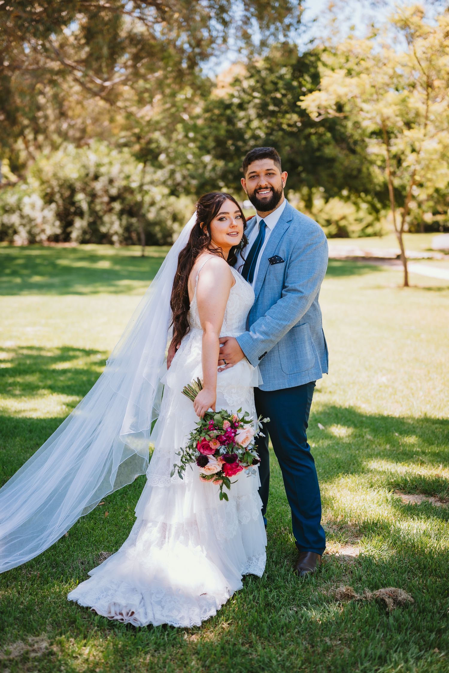 Newlywed couple poses on sunny lawn with flowing white veil and burgundy bouquet in outdoor wedding photo.