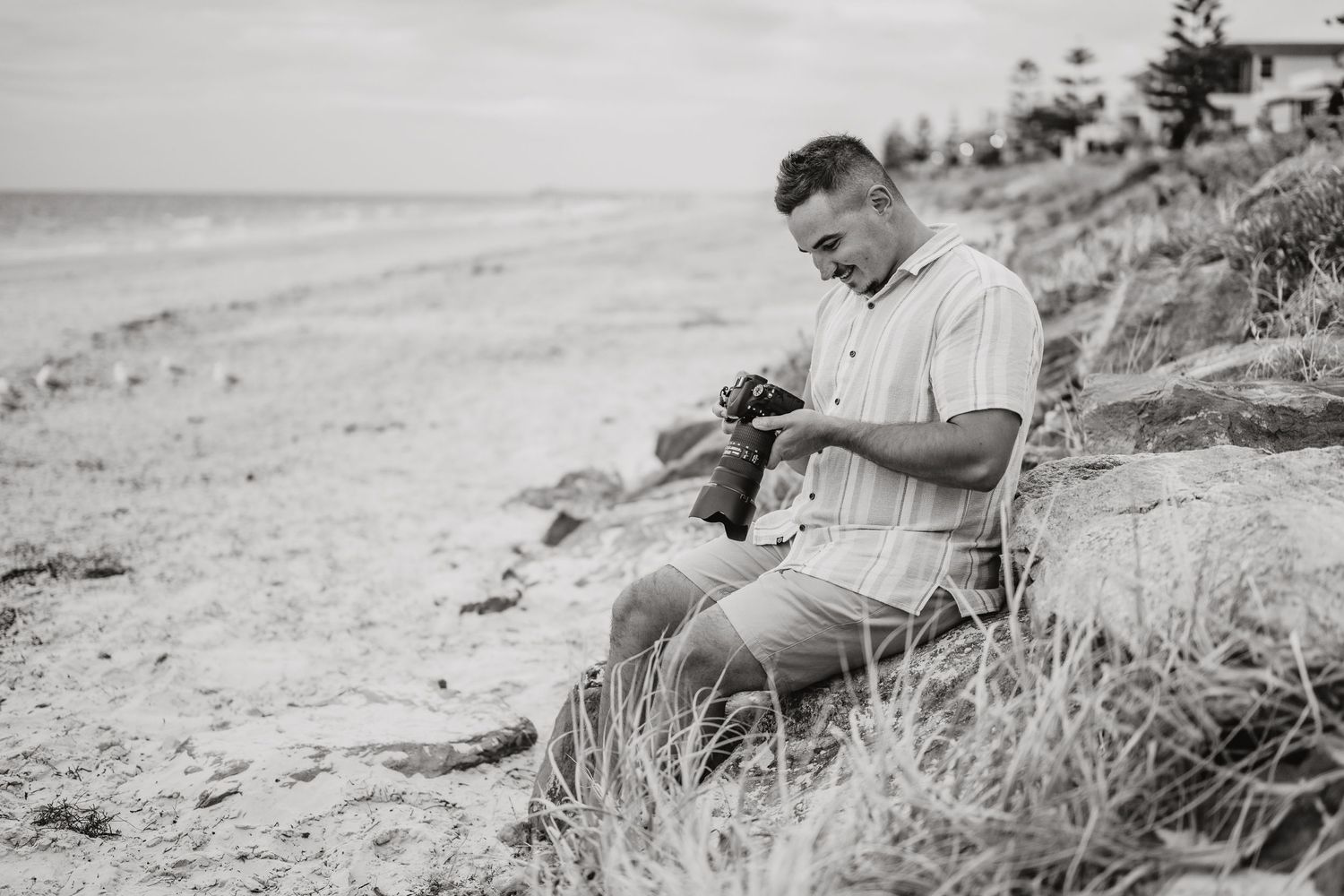A person in casual attire sits on a grassy beach slope reading with the ocean shore in the background during the 1950s.