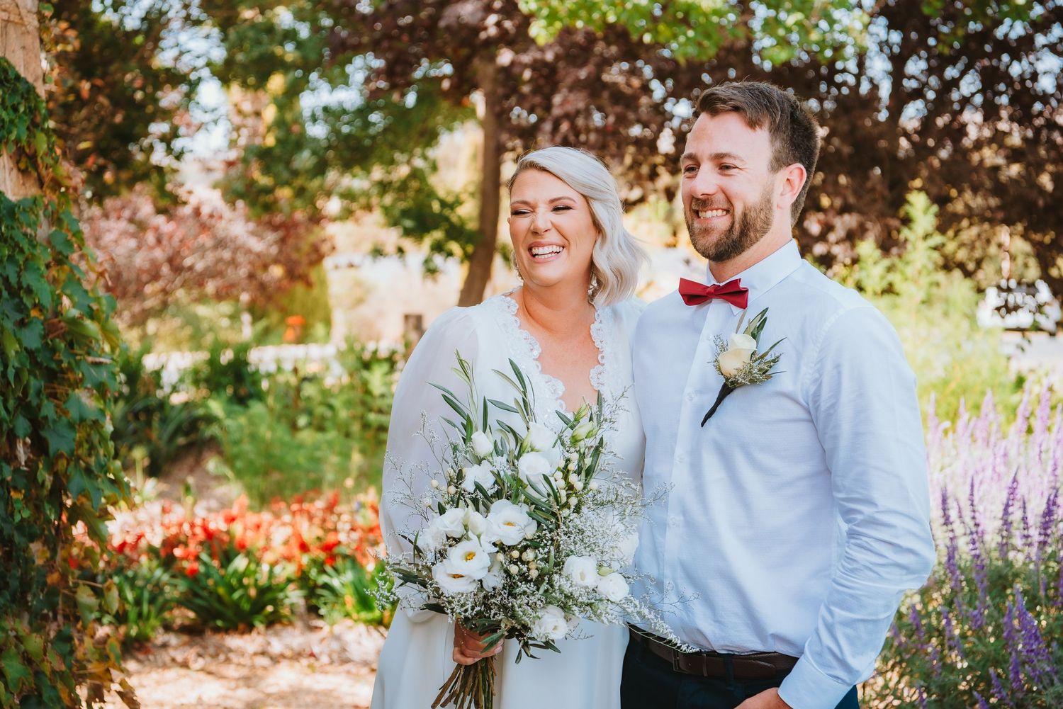 Happy couple shares a candid moment in a vibrant garden with blooming flowers and eucalyptus bouquet.