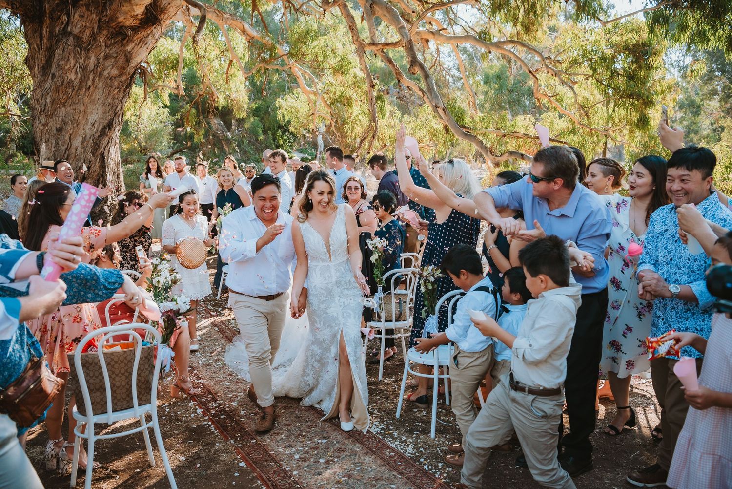 Wedding guests cheer from white chairs under trees as a couple walks down an outdoor aisle during a garden ceremony.