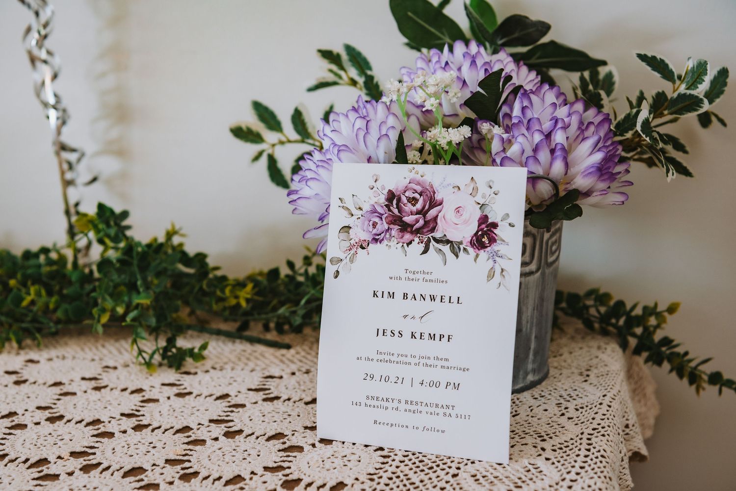 Wedding invitation card with burgundy floral design sits on lace tablecloth next to purple flowers in metal vase.