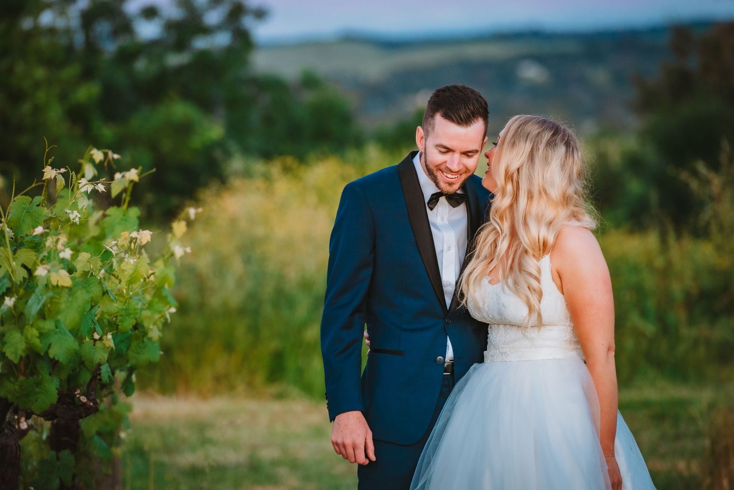 A couple shares an intimate moment together in a vineyard wearing formal wedding attire against a scenic landscape.