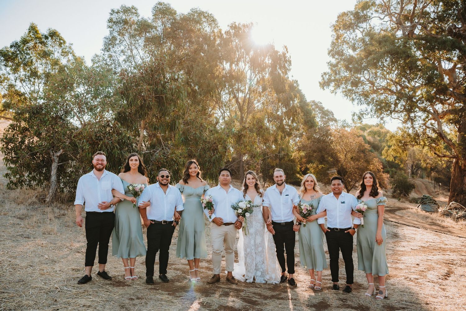 Wedding party group poses together on a sunlit dirt path with eucalyptus trees in the background at golden hour.