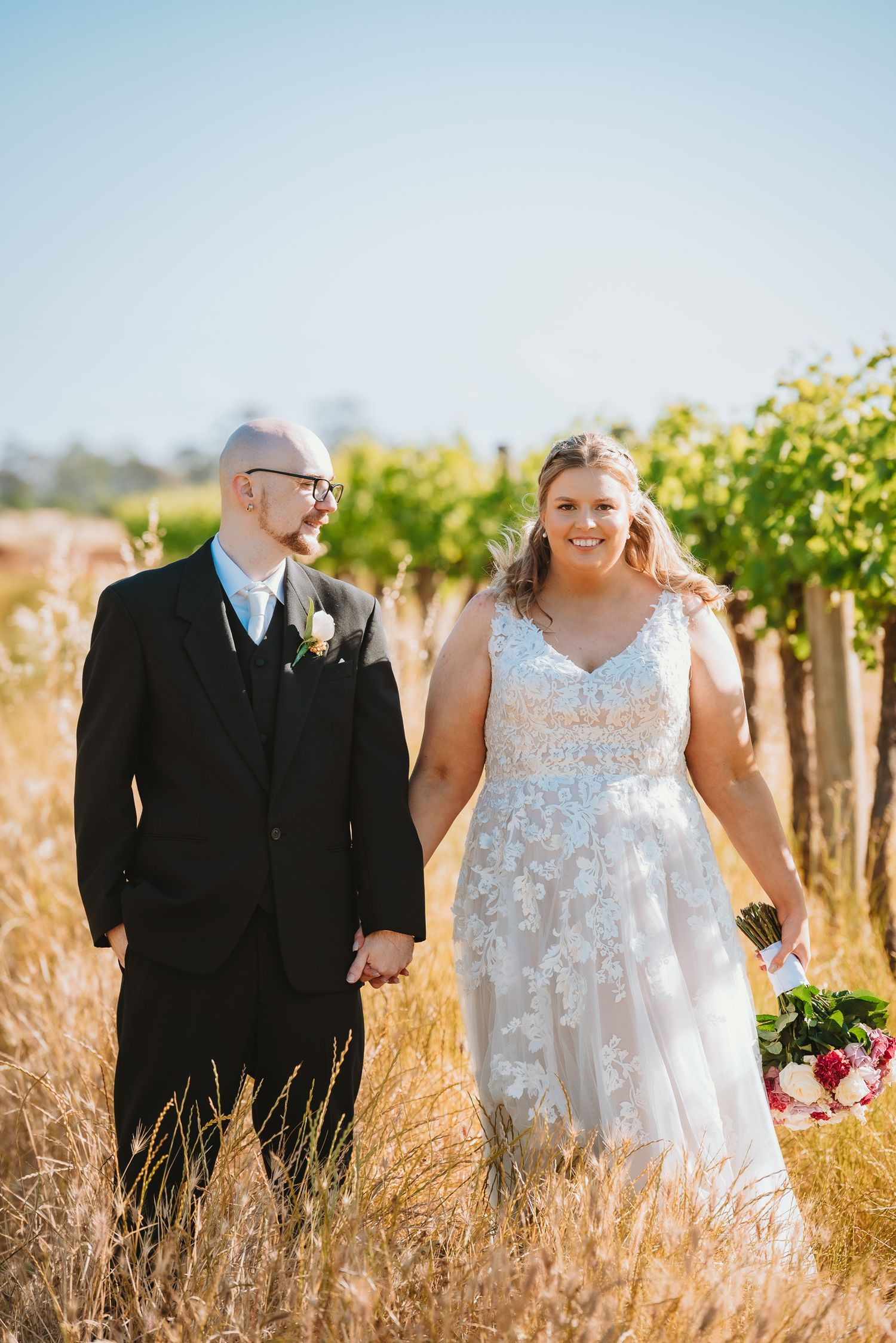 A couple walks hand in hand through a vineyard on their wedding day in golden sunlight.