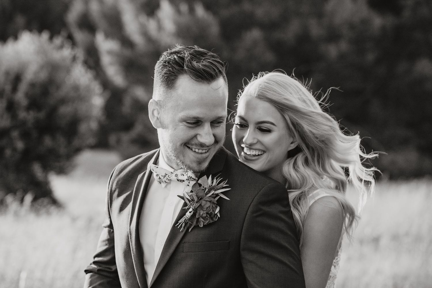 Black and white photo of a joyful couple laughing together outdoors, with the groom wearing a boutonniere.