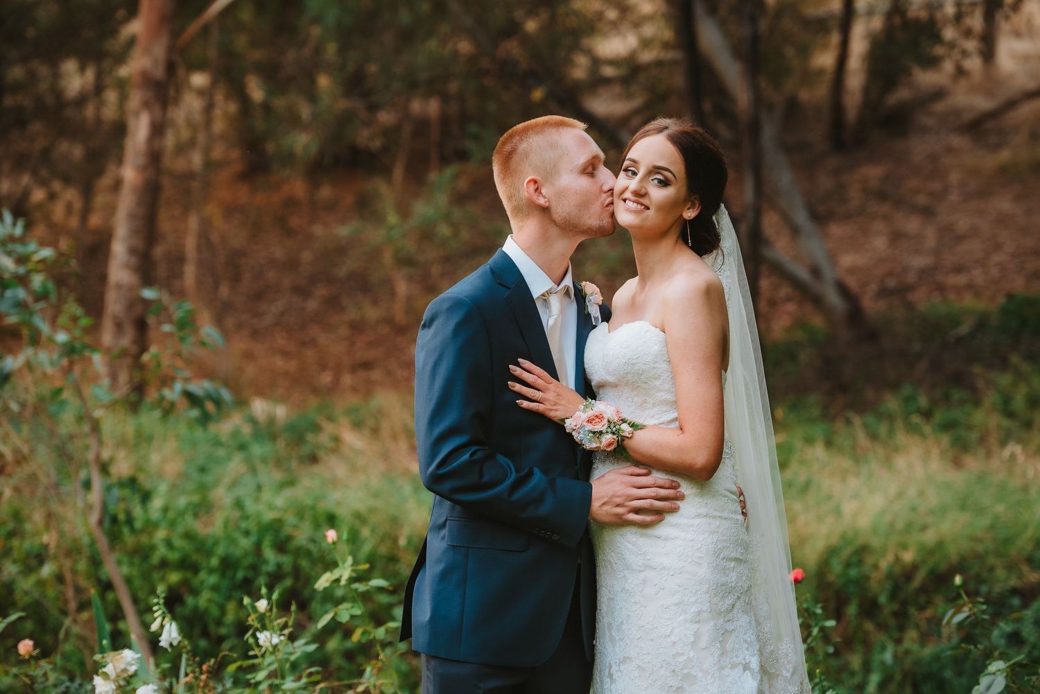 A bride in a lace dress and groom in navy suit share an intimate moment in a wooded outdoor setting.
