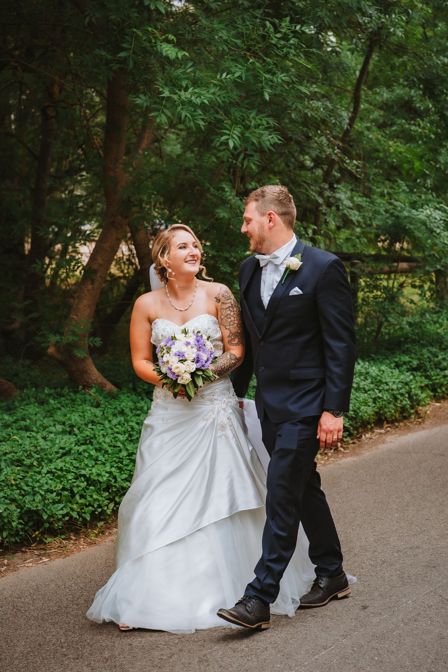 A couple shares a joyful moment while walking together on a tree-lined path, with purple bouquet and formal attire.
