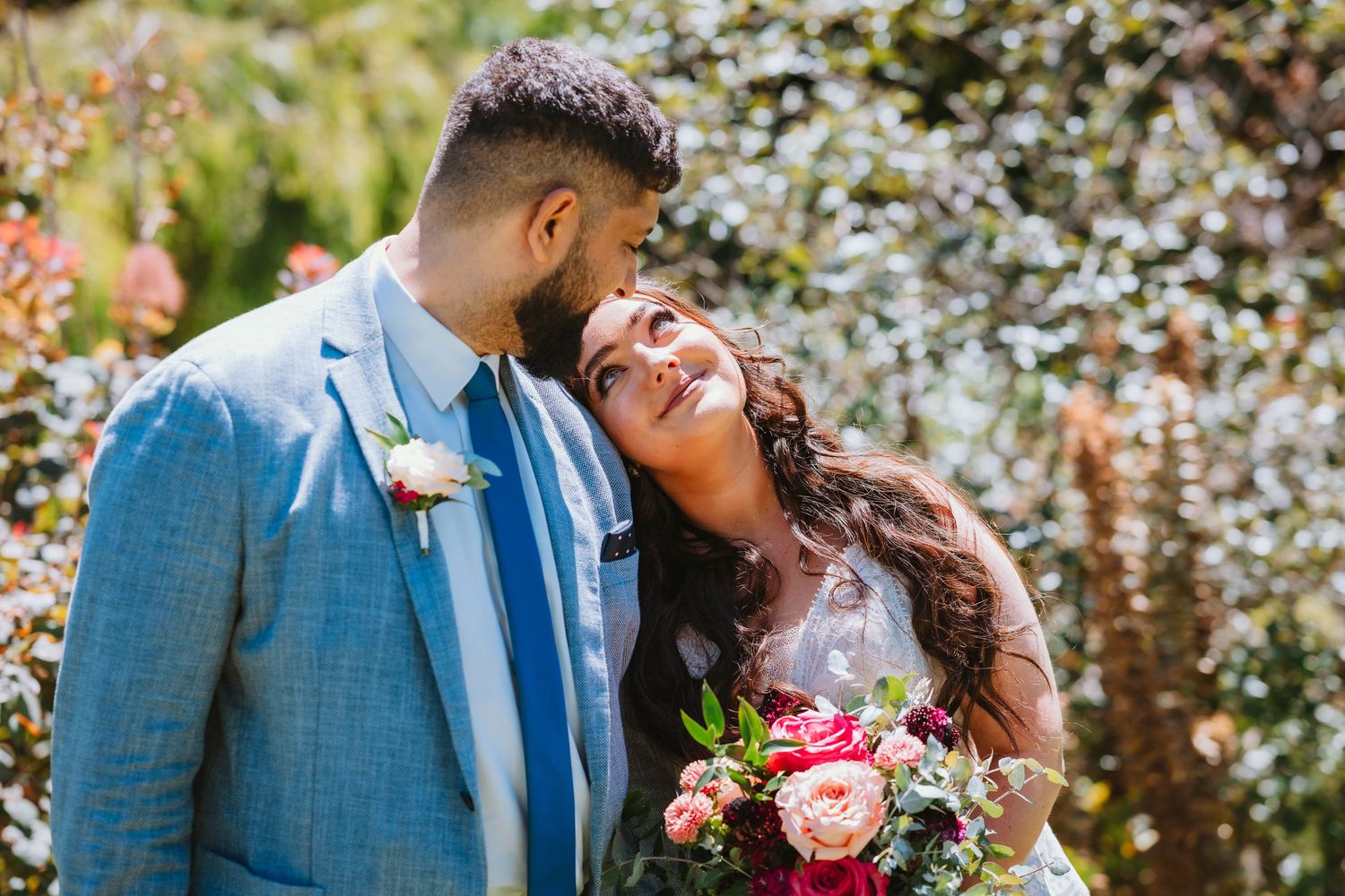 Couple shares a joyful moment at an outdoor wedding celebration with flowers and sunlit garden in background.