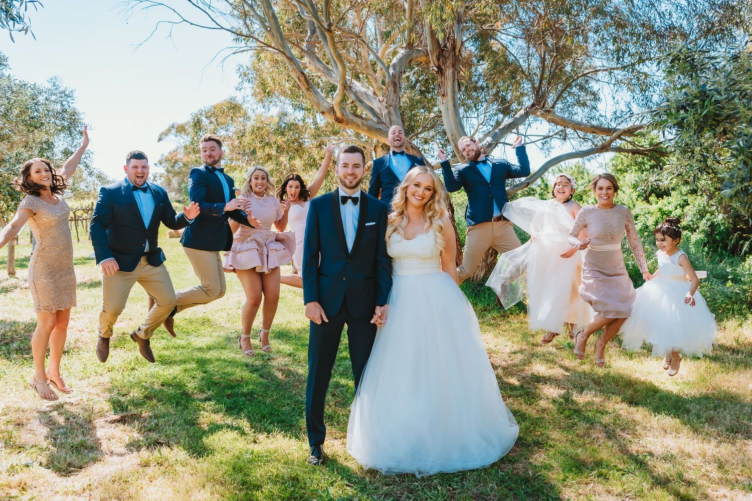 A wedding party leaps joyfully into the air on a sunny hillside with trees in the background.