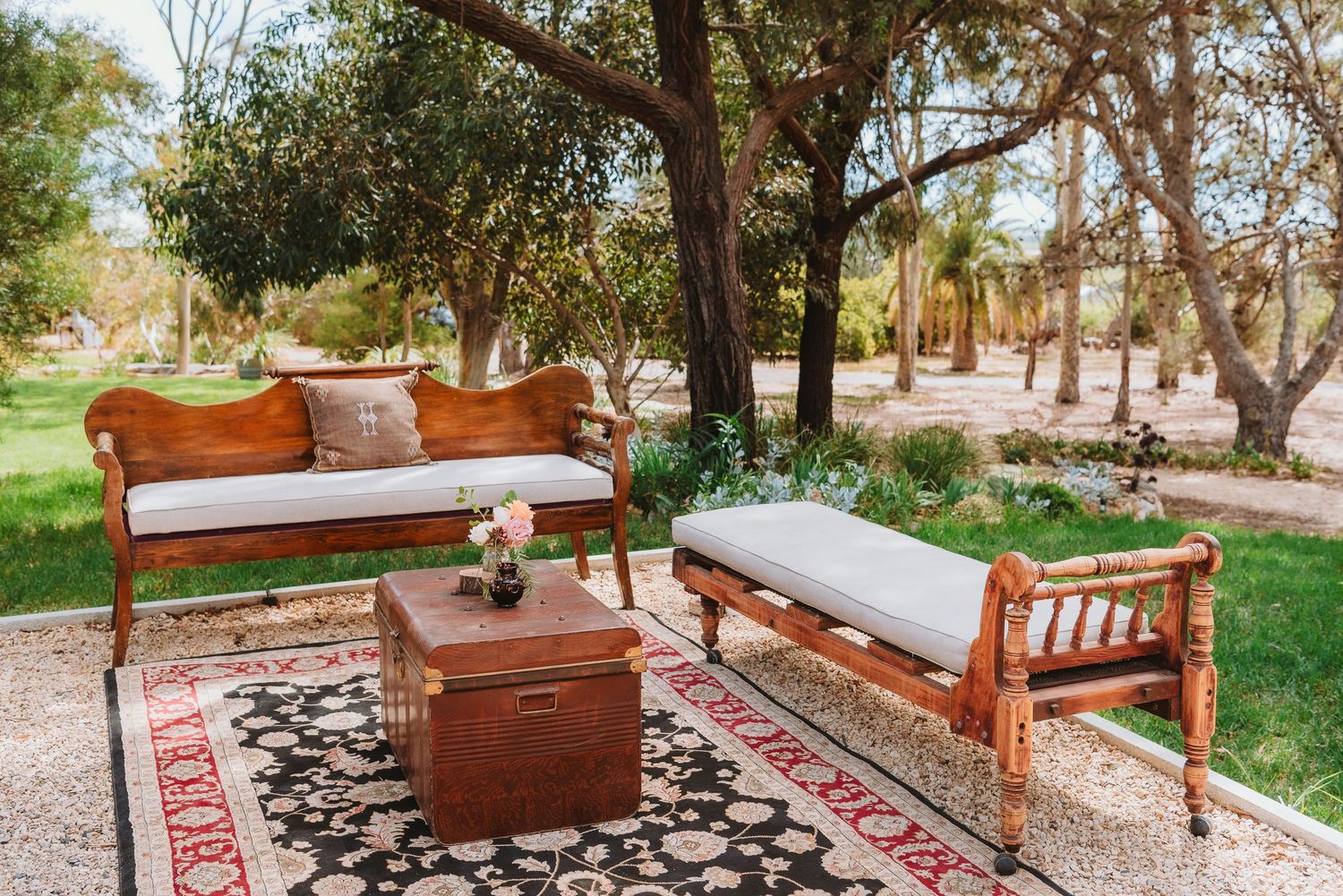 Vintage wooden seating arrangement with decorative rugs and cushions in an outdoor garden setting under trees.