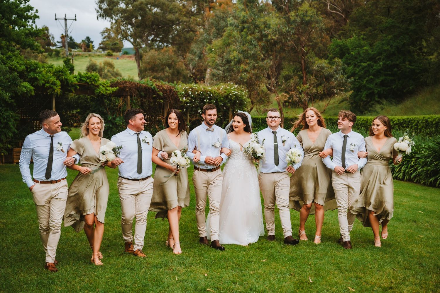 Wedding party in light beige and blue attire walking together on a lush green lawn during an outdoor celebration.