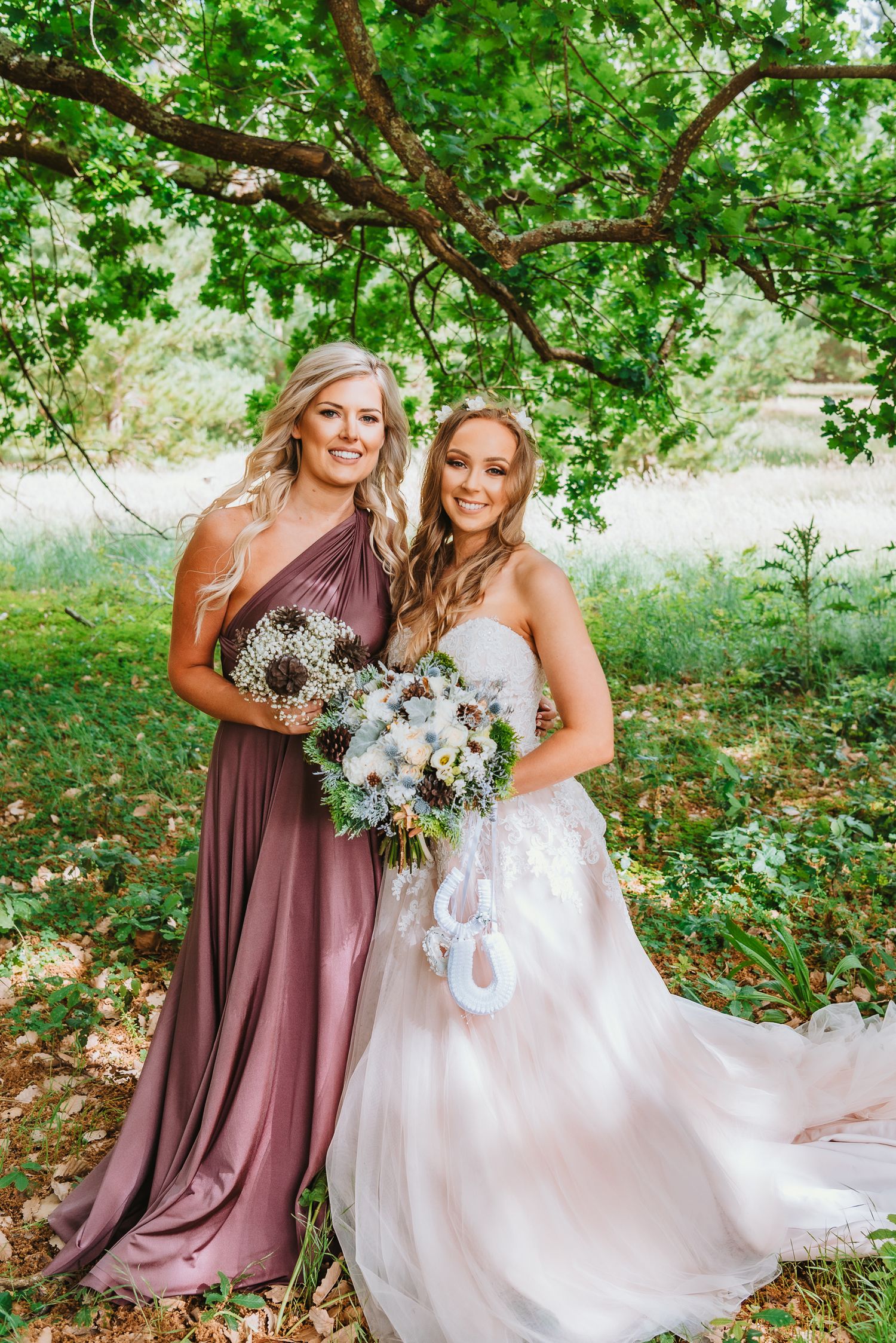 Two women stand under a tree branch, one wearing a mauve bridesmaid dress and another in a flowing white wedding gown.