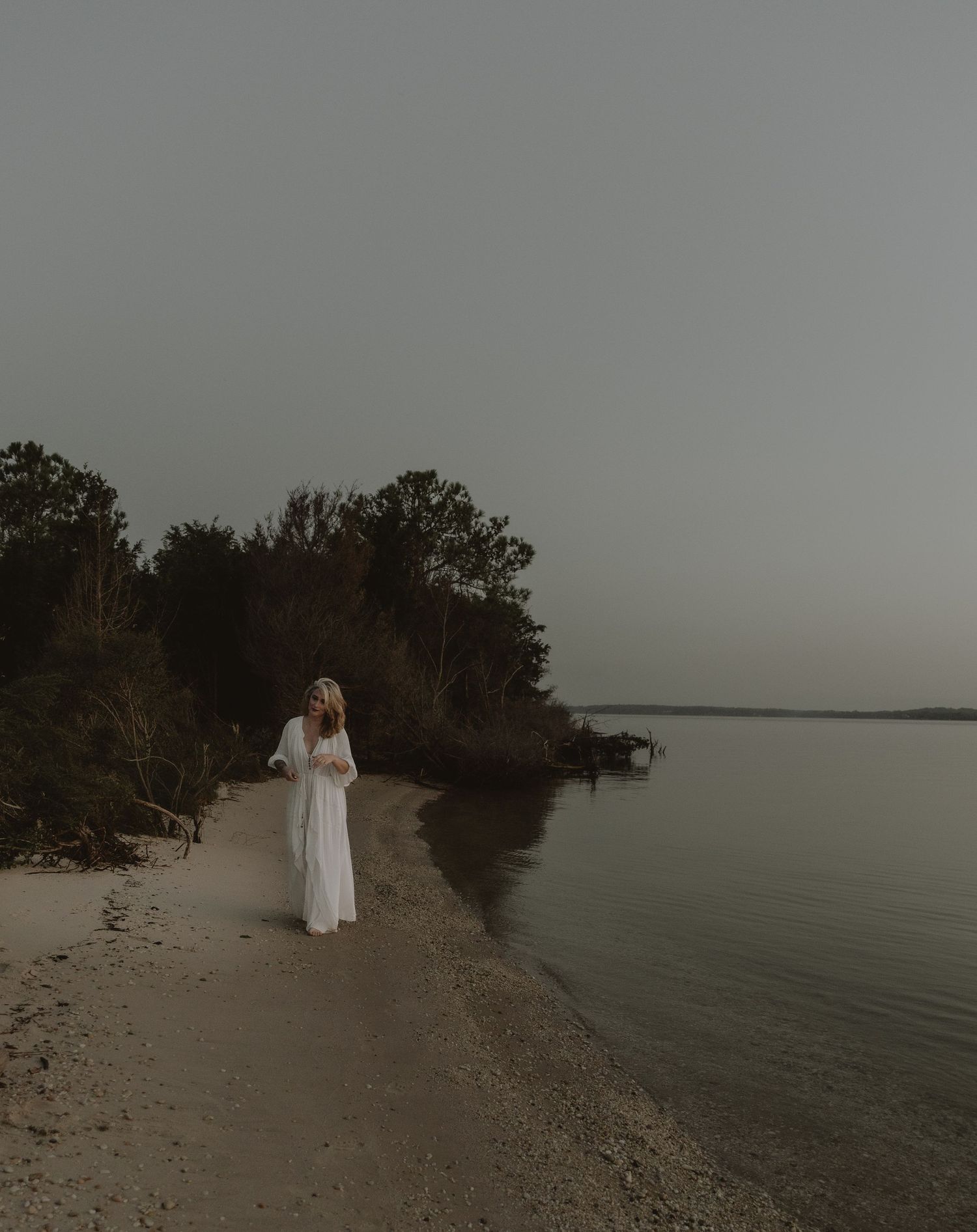 Ethereal figure in white dress standing on shoreline surrounded by trees at twilight.