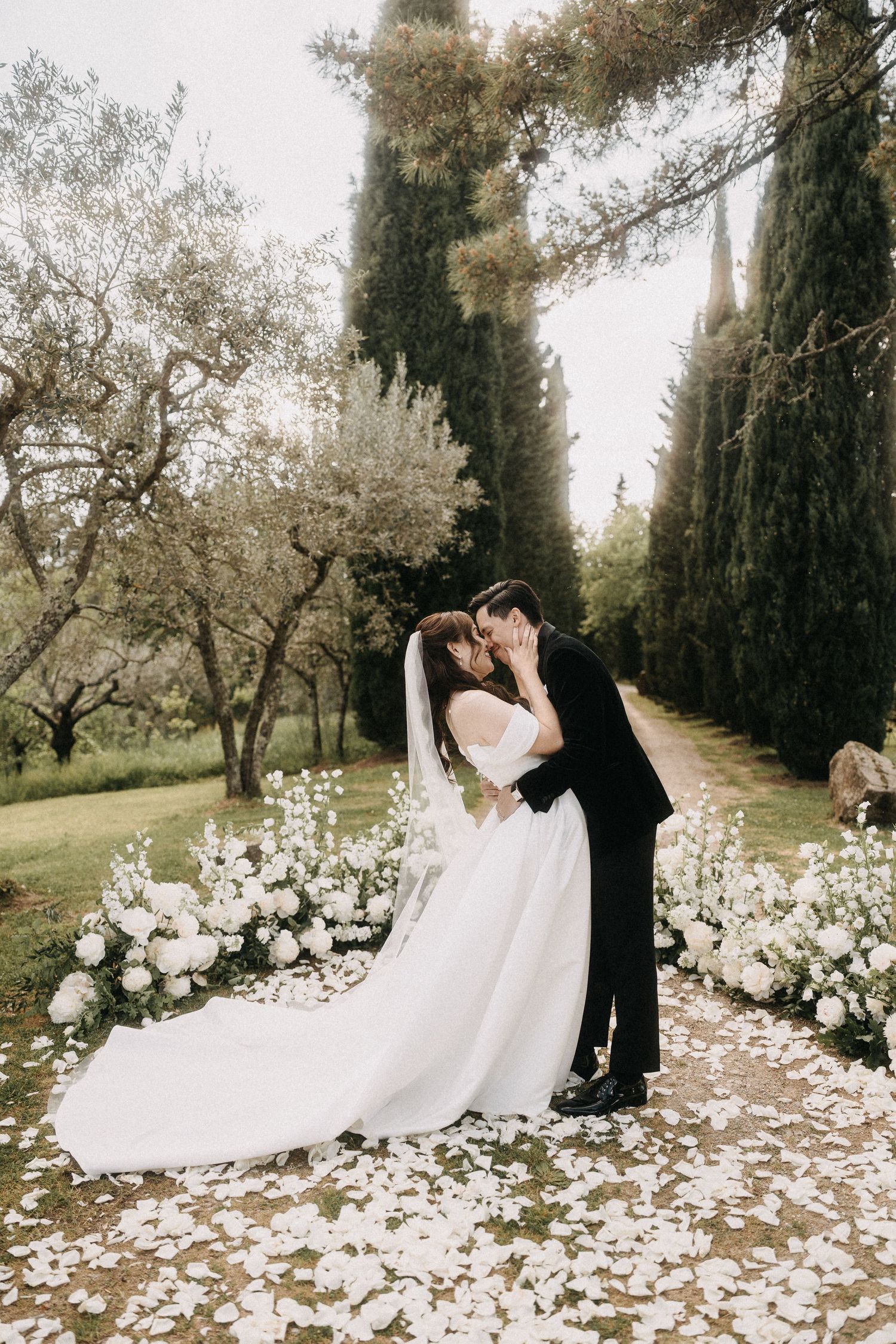 A romantic wedding couple shares a kiss on a garden path lined with cypress trees and scattered white flower petals.