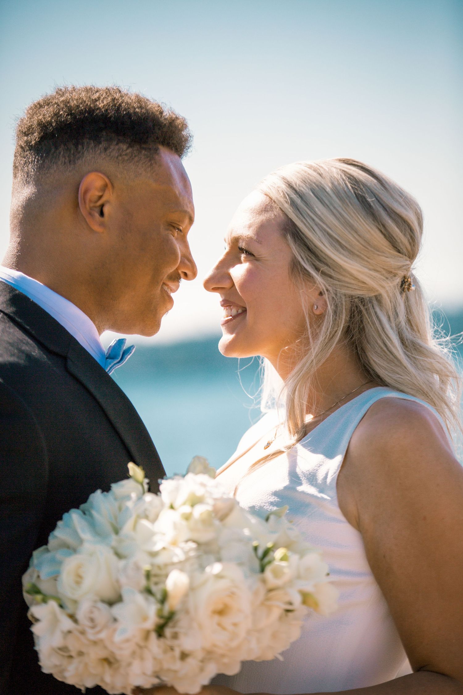 Romantic newlywed couple sharing intimate moments by the ocean with white floral bouquet.