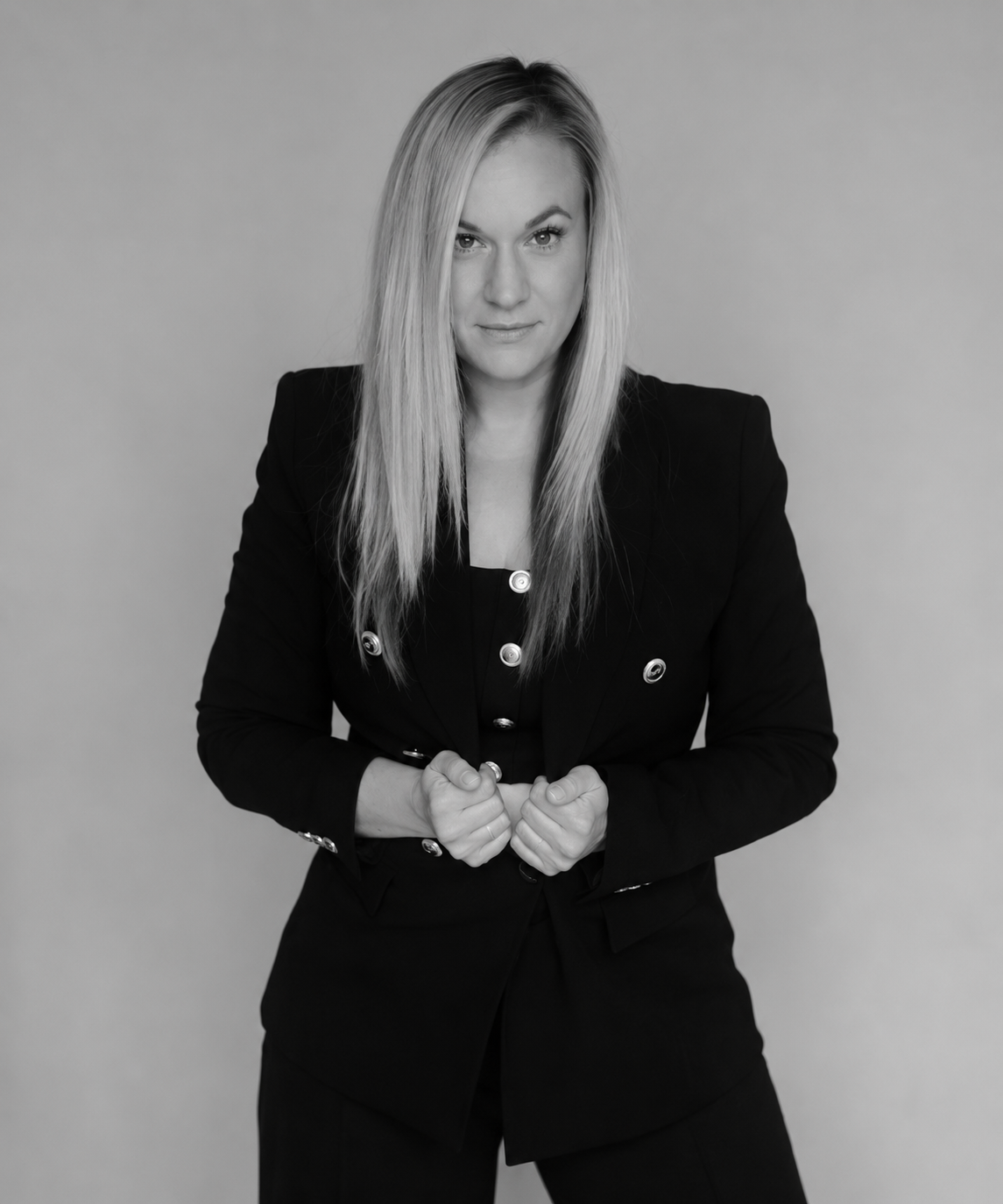 A blonde woman in a black blazer suit poses confidently with clasped hands in a professional black and white portrait.