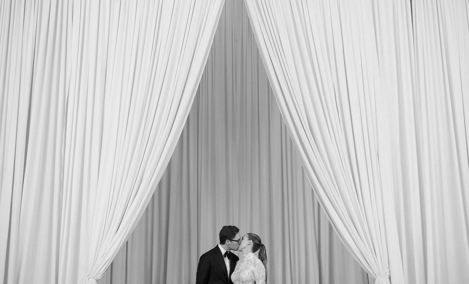 Black and white photo of a bride and groom sharing a kiss framed by elegant white draping curtains.