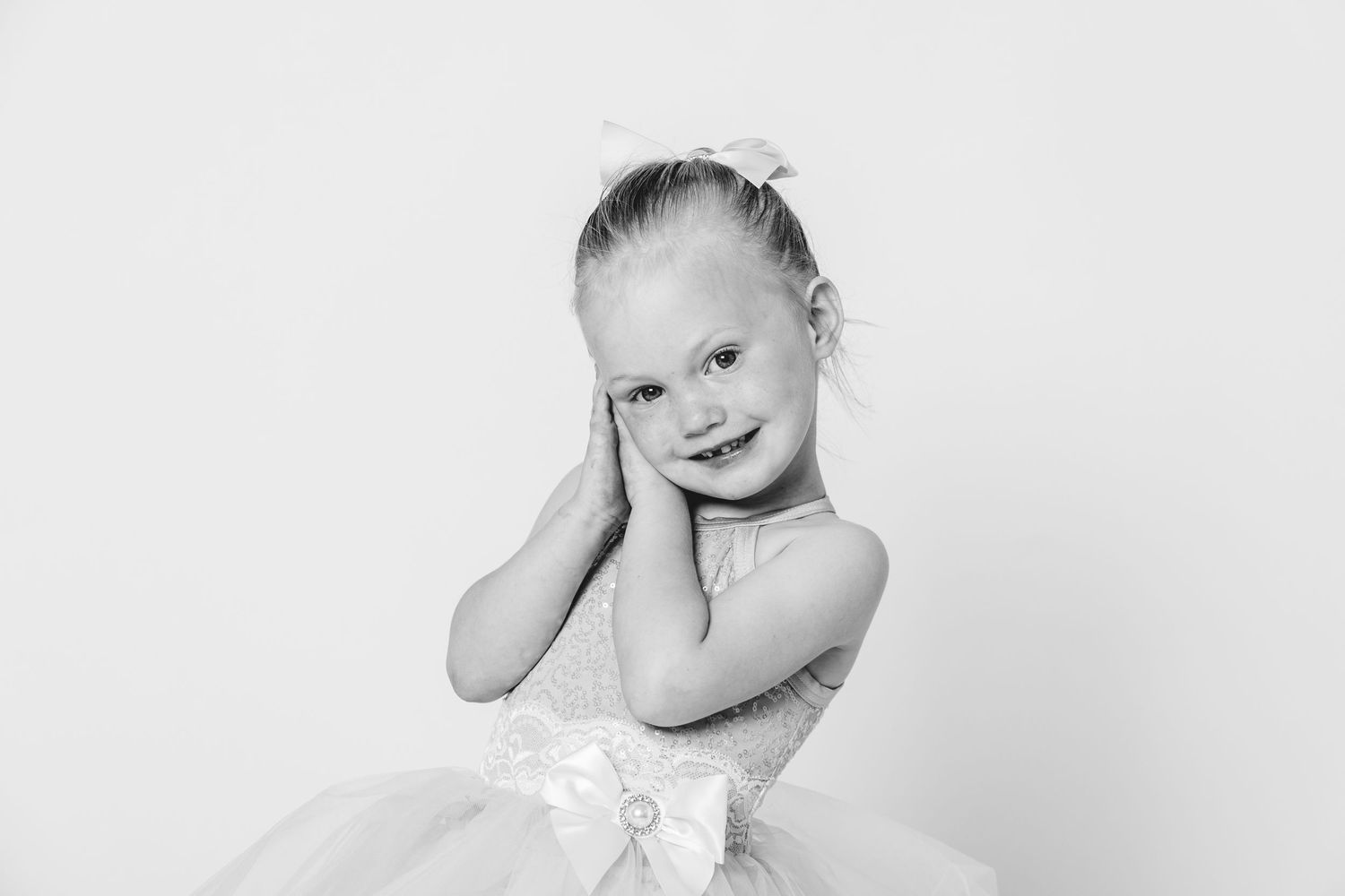 Black and white portrait of a smiling little girl in a tutu dress with a bow, hands on cheeks, white background.