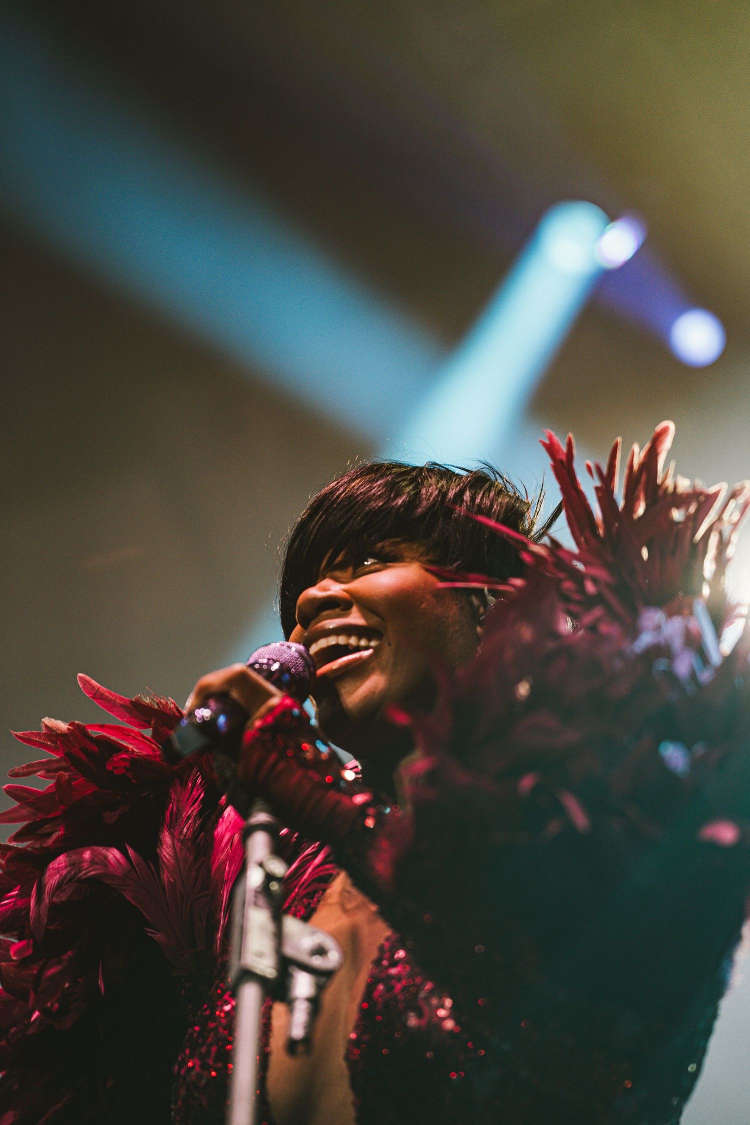 A joyful Black female singer performs on stage wearing a dramatic red feathered costume under blue spotlights.