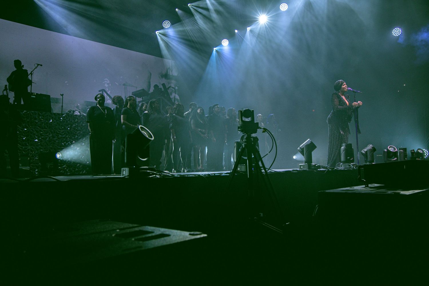 Live concert stage with performers under dramatic blue and white spotlights in a large dark venue.