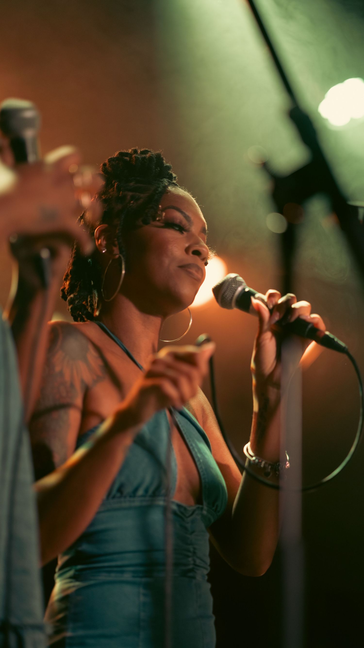 A woman singer performs on stage holding a microphone under warm stage lights wearing a denim top with hoop earrings.