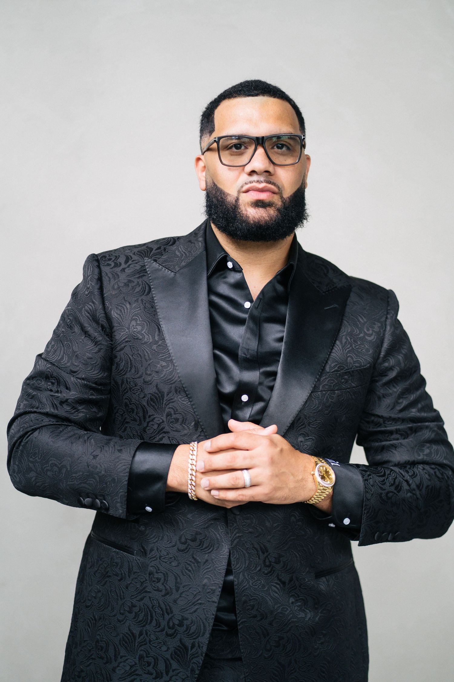 Studio portrait of person in black patterned suit with glasses and jewelry.