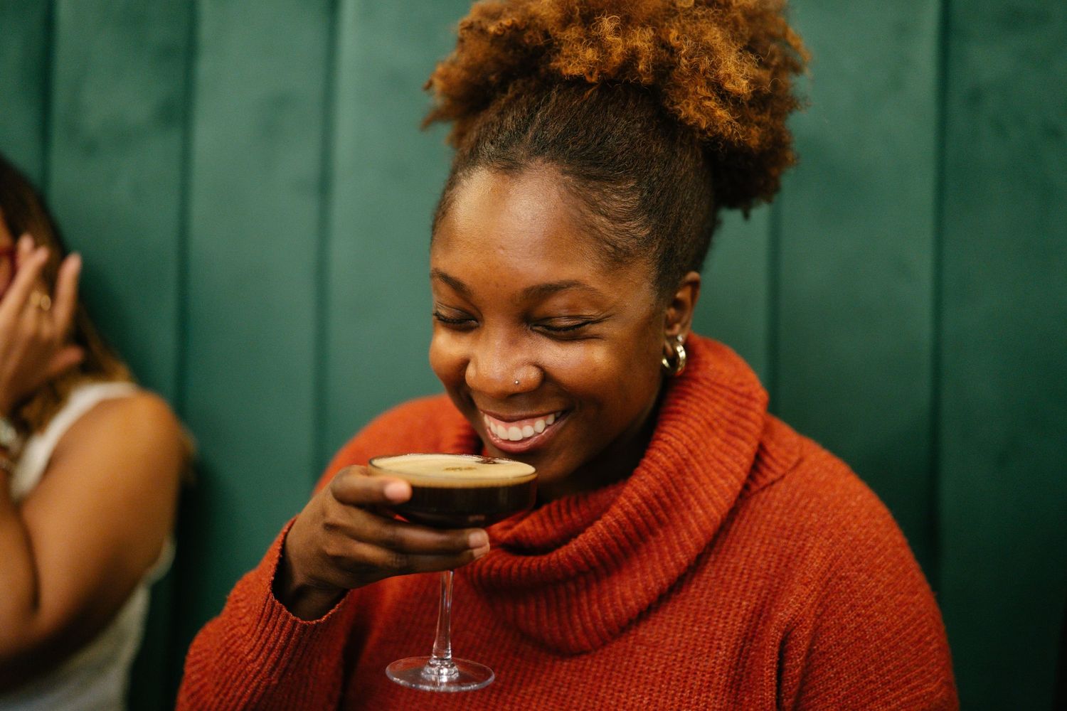 Smiling Black woman in orange turtleneck sweater enjoying a cocktail at a bar.