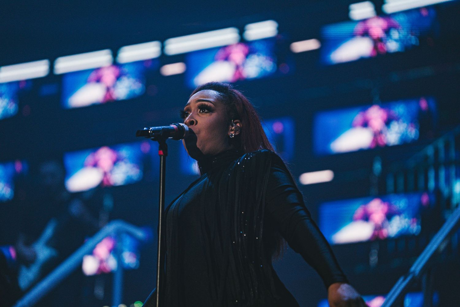Female singer performing on stage in black outfit with microphone, dramatic blue lighting and LED screens behind her.