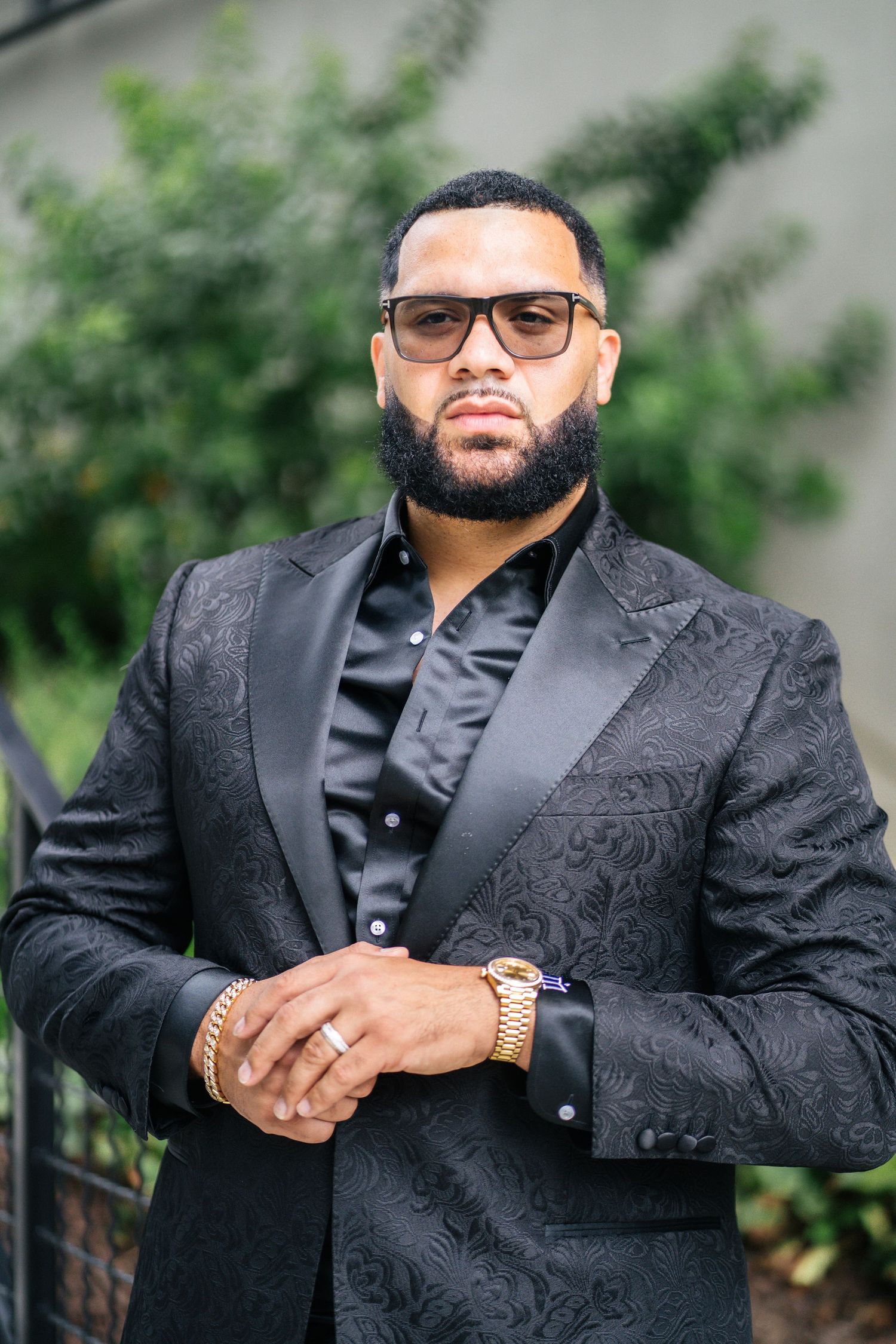 Outdoor portrait of person in black formal suit against green foliage background.