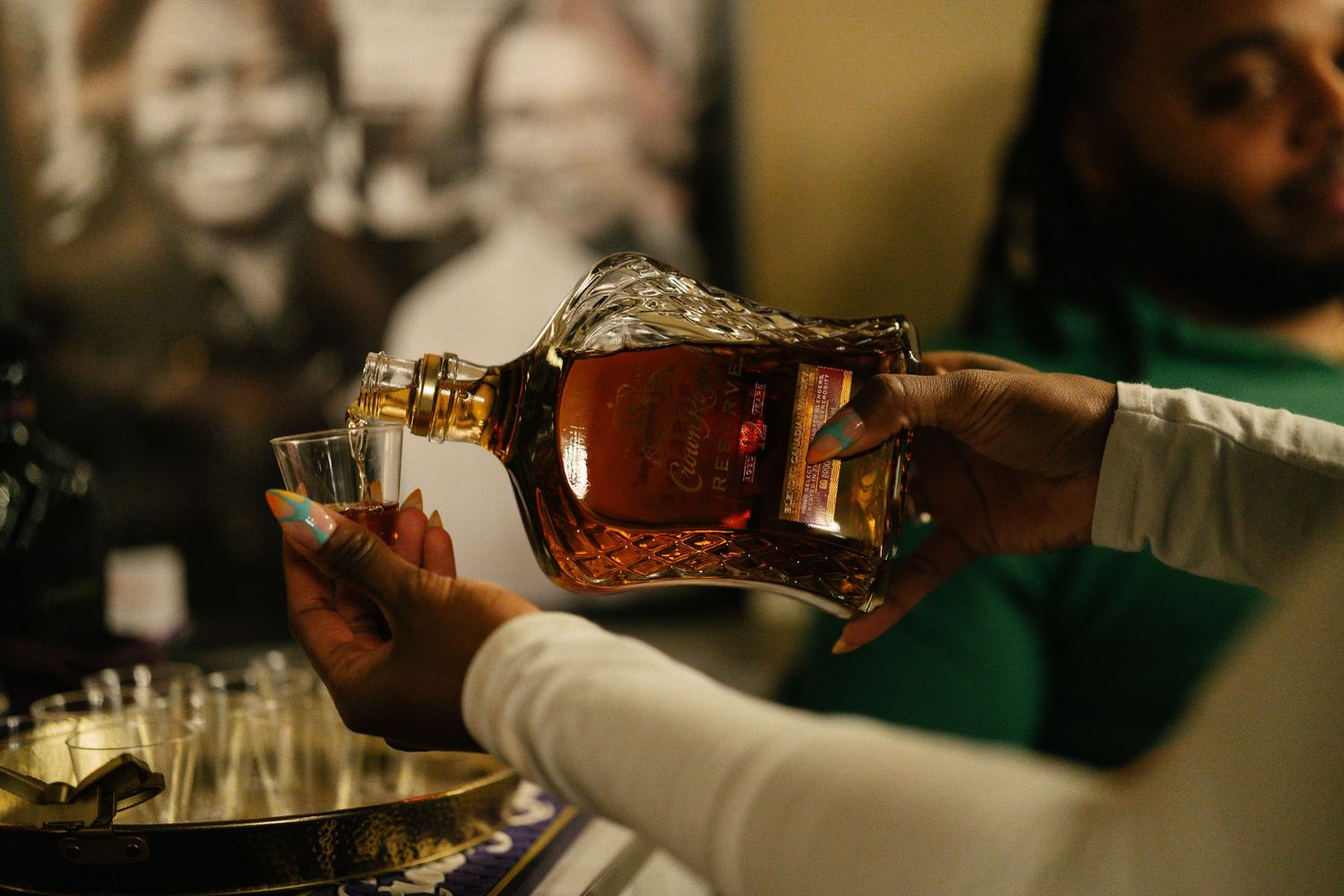 Person pouring amber whiskey from decorative crystal decanter into shot glass at a bar event.