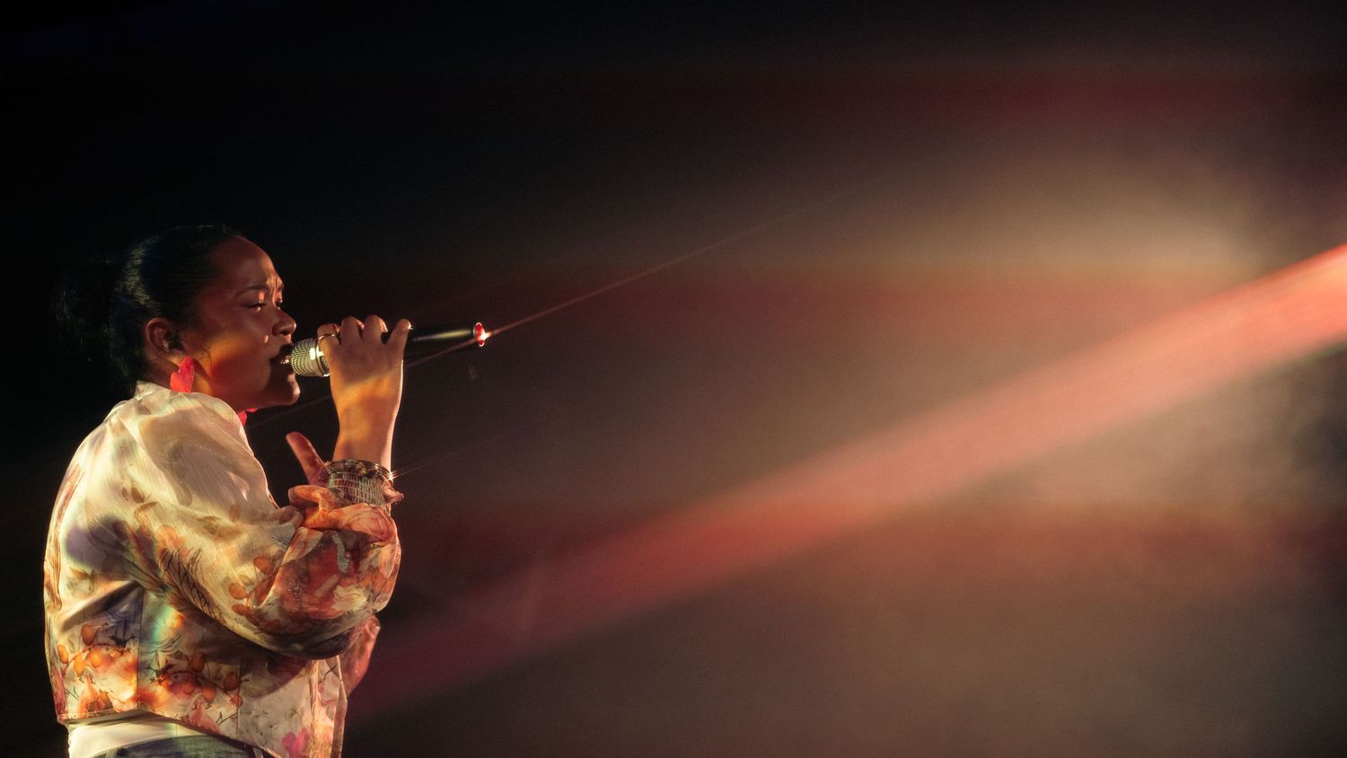 Singer performing on stage with dramatic red and orange light beams cutting through dark atmosphere.